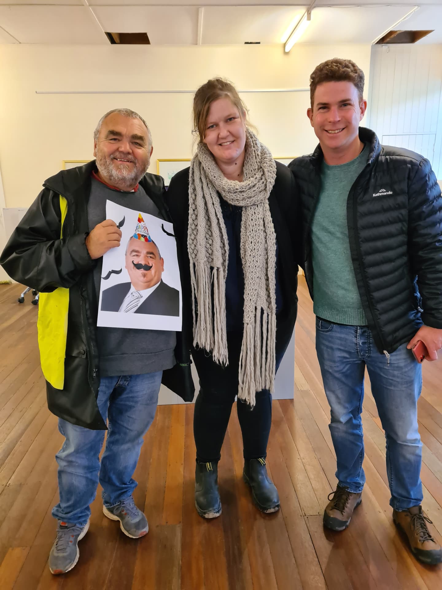 Mike Haywood with two colleagues in the program site office, holding a birthday poster made by the team
