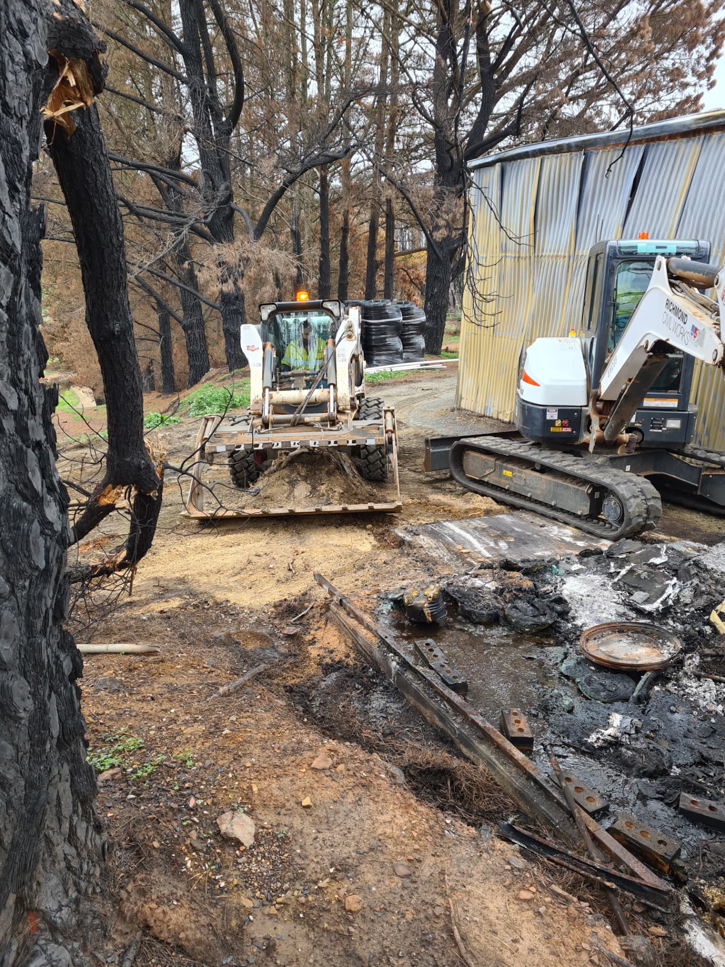 Skid-steer loader and tracked excavator clearing debris between burnt trees beside a fire-damaged shed